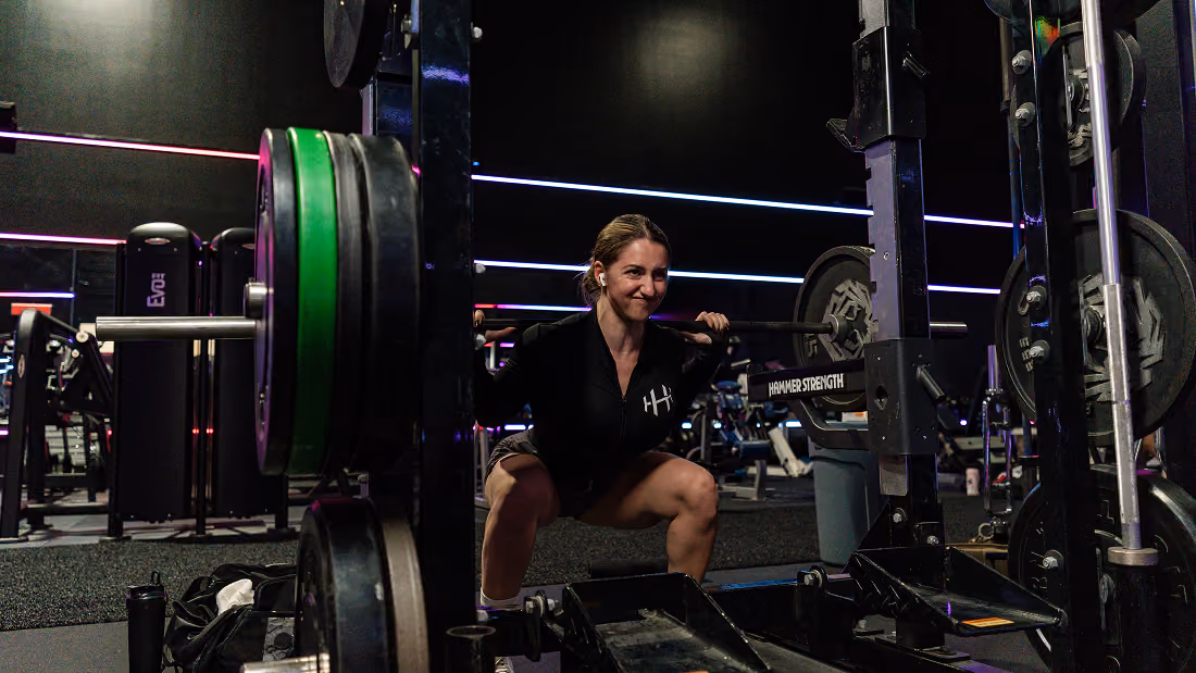 Woman performing weighted barbell squat in a gym with neon lights and fitness equipment.
