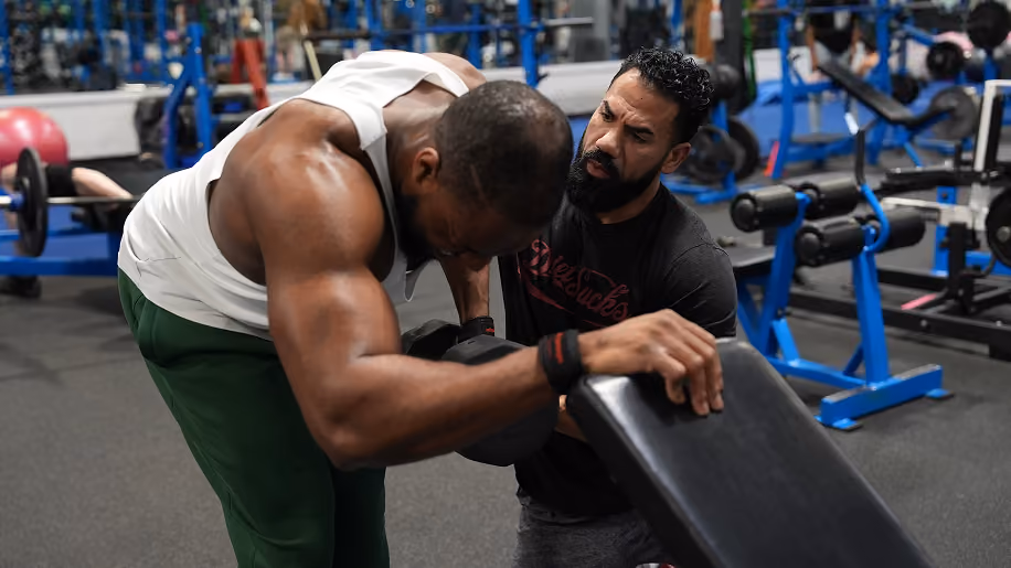 A muscular man in white tank top exercises with a bench while a trainer watches closely in a gym.