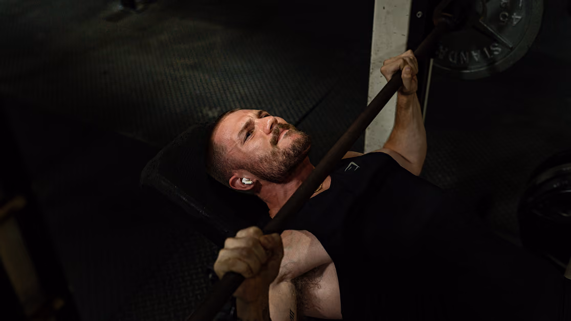 Man intensely lifting a barbell while lying on a bench in a dimly lit gym.