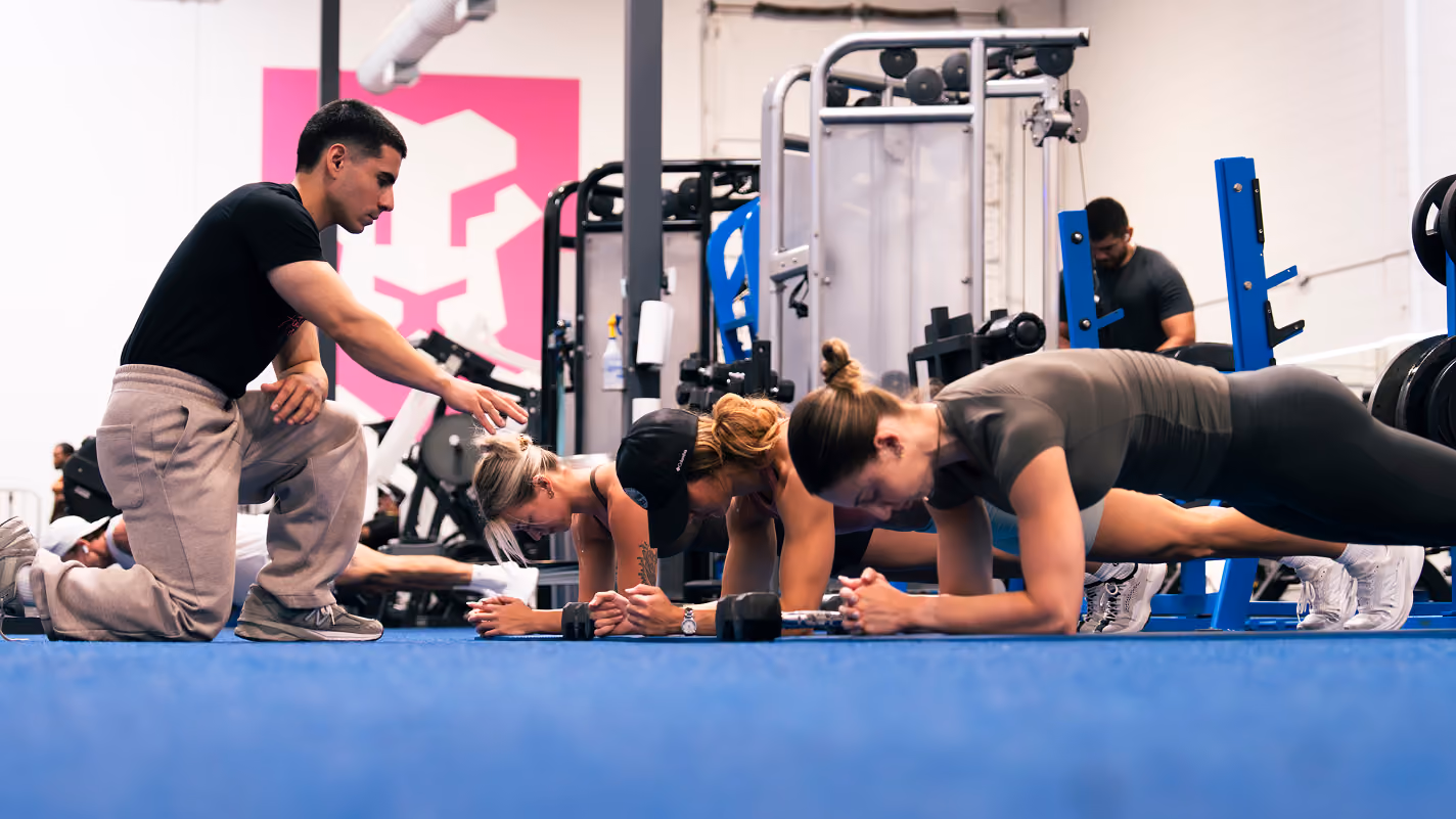 Three people in a gym doing planks on a blue mat while a trainer kneels beside them giving guidance.