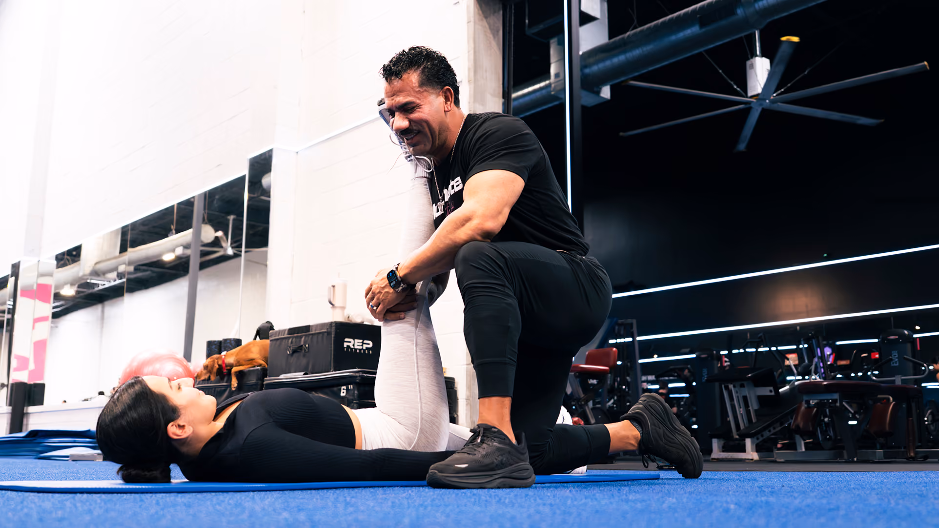 Personal trainer assisting a woman lying on a mat with a leg stretch in a gym.