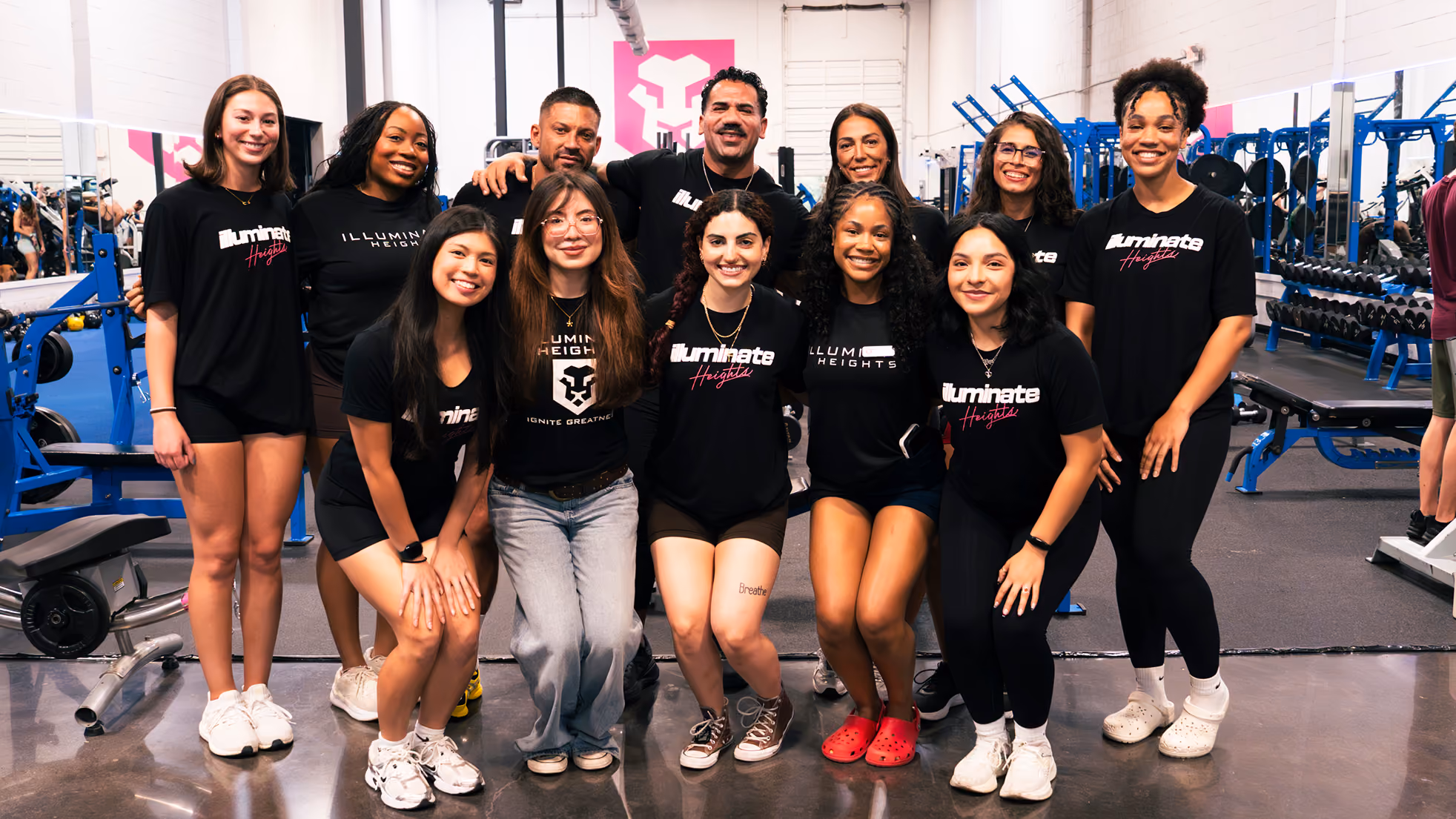 Group of smiling young adults in black Illuminate Heights shirts posing in a gym with weight equipment.