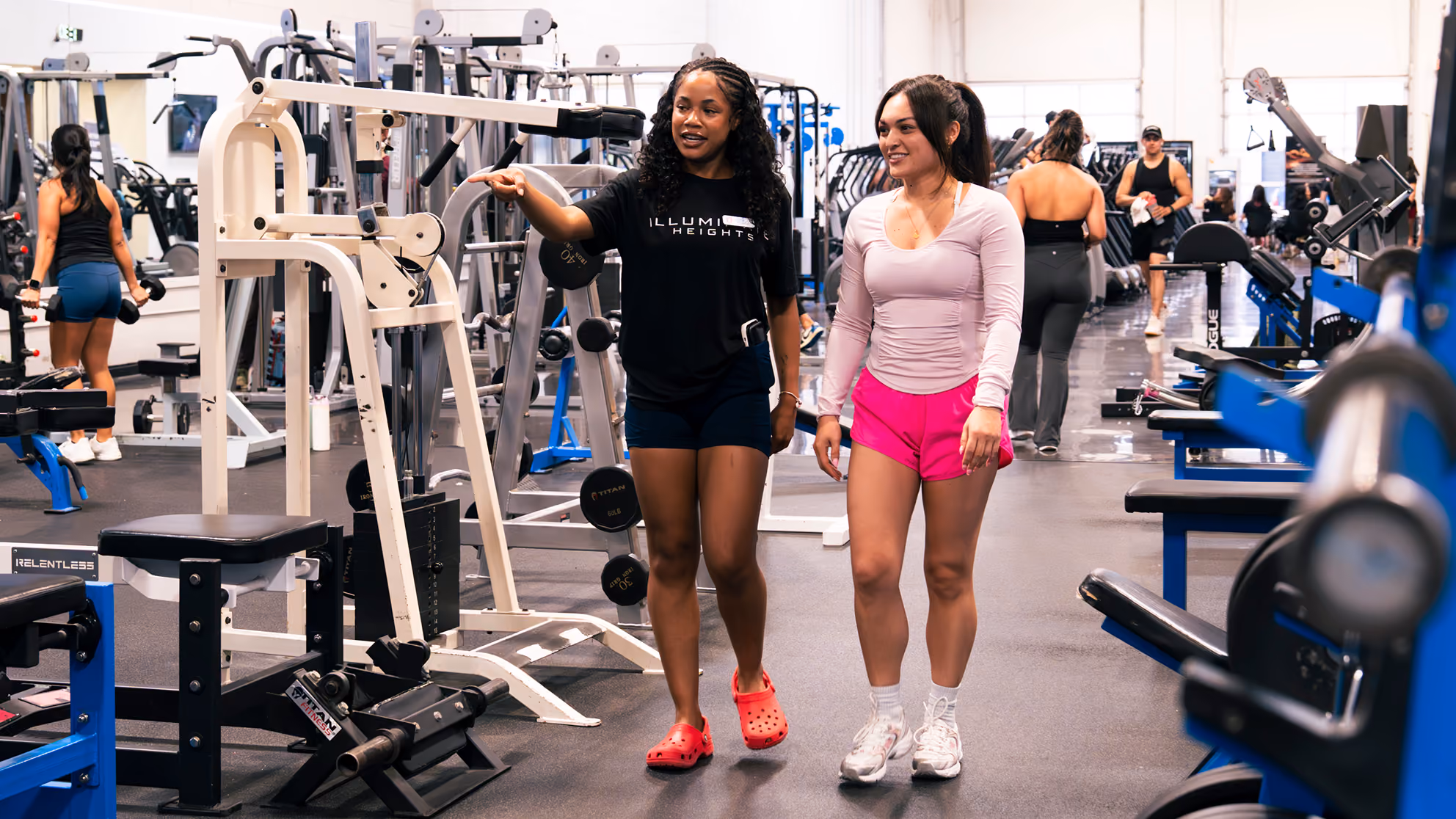 Two women walking and talking inside a gym surrounded by workout equipment and other people training.