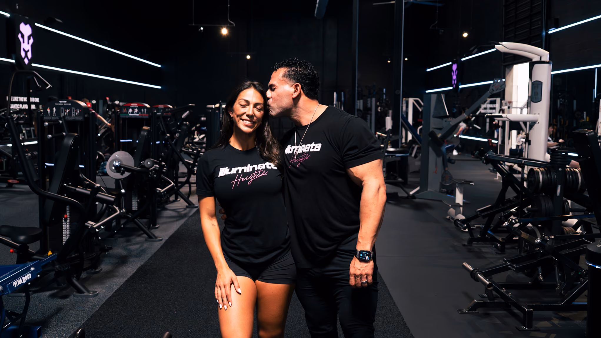 Nate Bran kisses Jourdan Bran on cheek in gym while both wear matching Illuminate Heights shirts.
