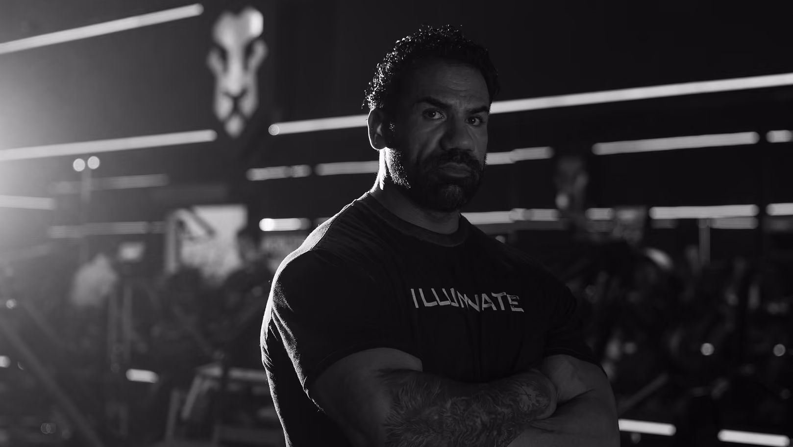 Muscular man with tattoos stands with crossed arms in a gym, lit dramatically in black and white.