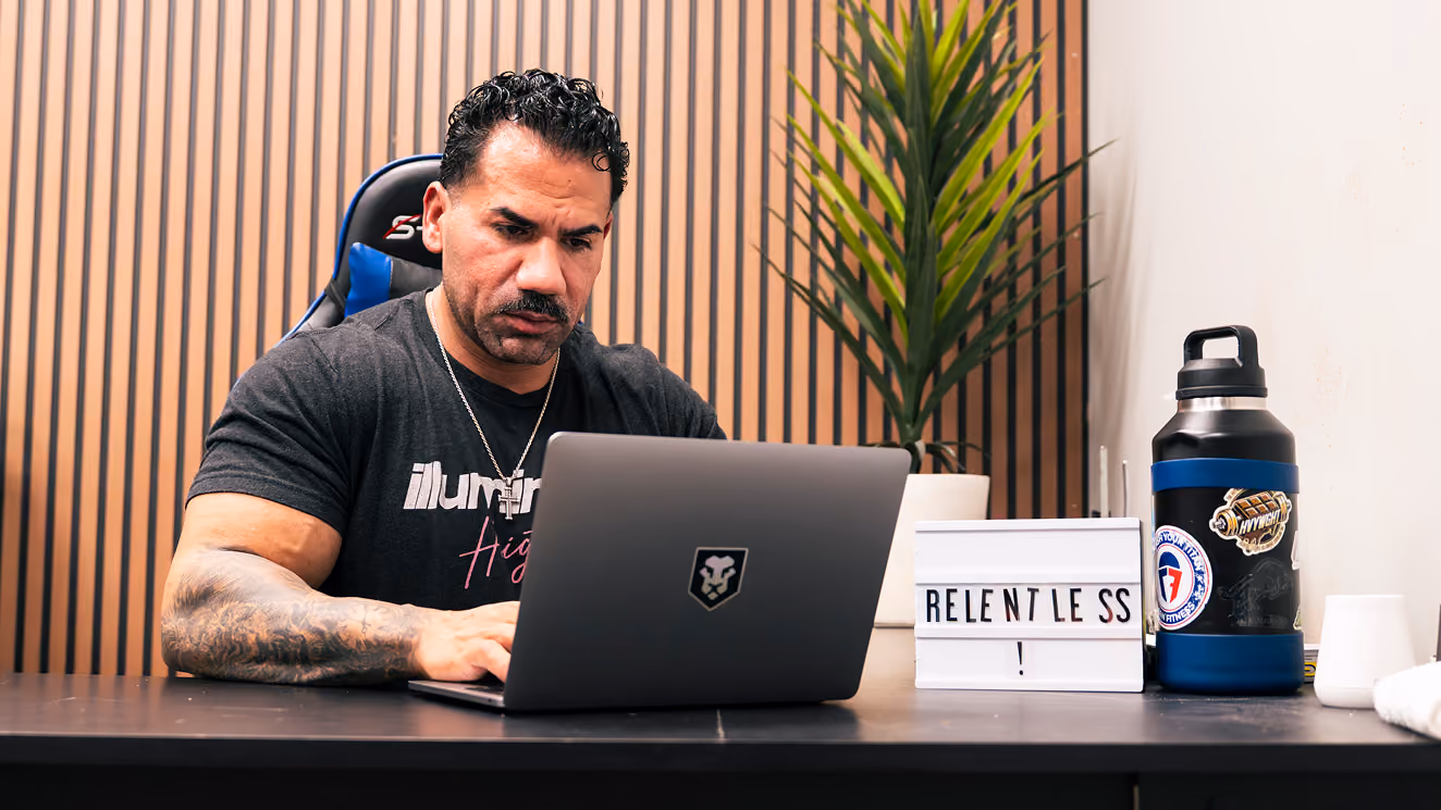 Man with a tattooed arm working on laptop at a desk with a 'RELENTLESS!' sign and water bottle.