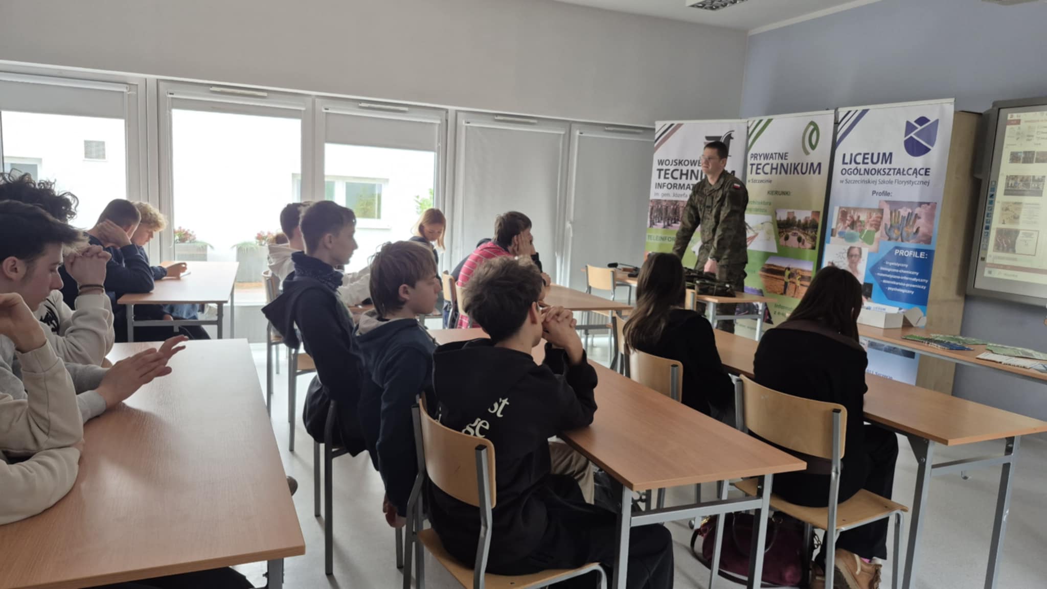 Uczniowie siedzą w klasie, słuchając wykładu żołnierza przy biurku i tablicy z banerami edukacyjnymi.