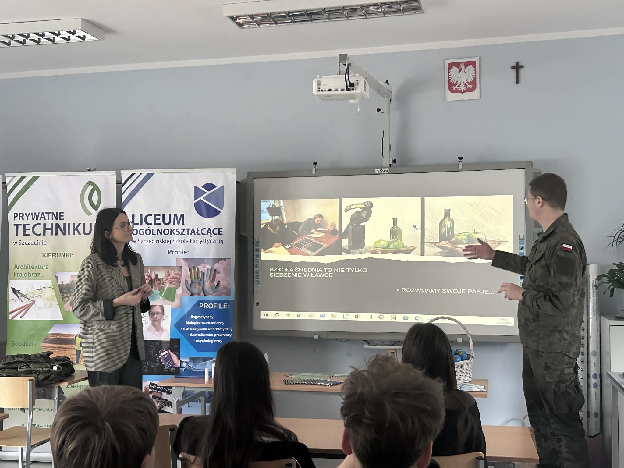 Kobieta i żołnierz prowadzą prezentację dla uczniów w klasie z rzutnikiem i plakatami edukacyjnymi.