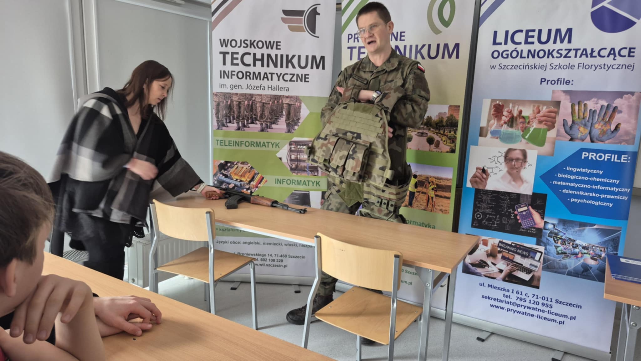 Kobieta sięga po karabin na stoliku, obok stoi mężczyzna w mundurze z kamizelką kuloodporną.