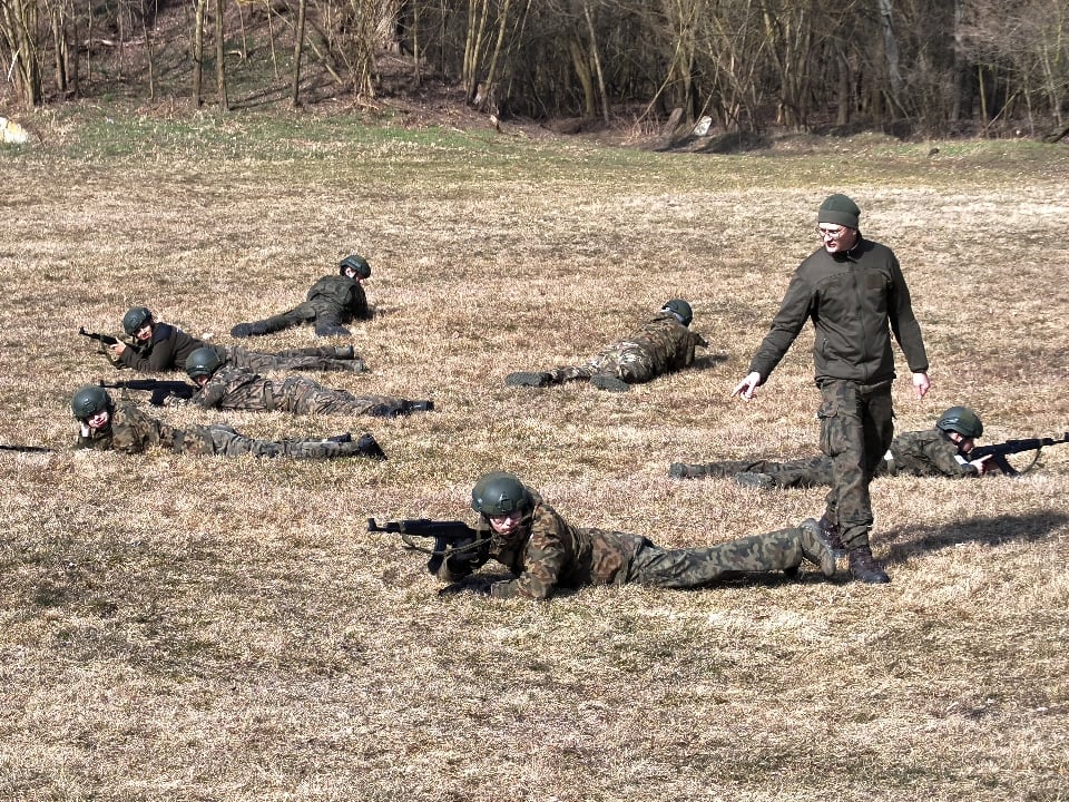 Grupa żołnierzy leżących na trawie podczas ćwiczeń strzeleckich, z instruktorem stojącym obok.