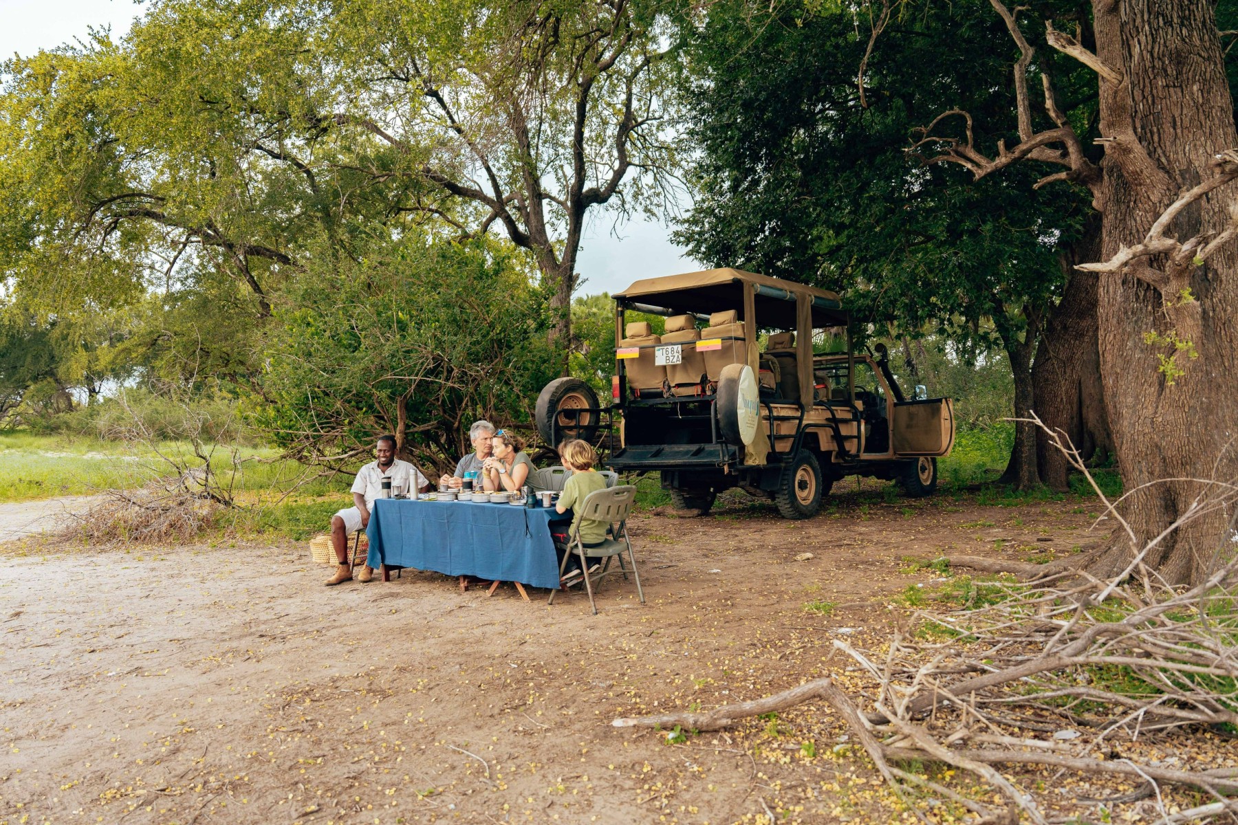 Travelers dining beside a safari vehicle beneath trees in East Africa