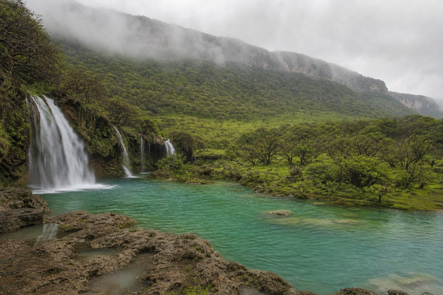 A waterfall spilling into a green pool below the monsoon-green hills above Salalah