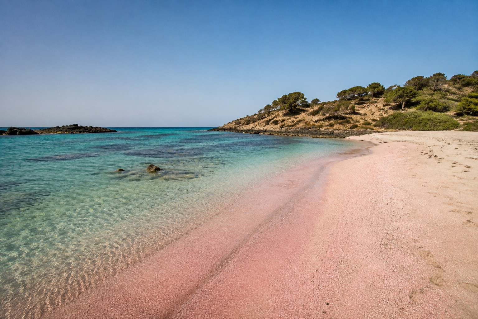 Empty pink tinged sand curving into shallow turquoise water at Elafonisi on the west coast of Crete