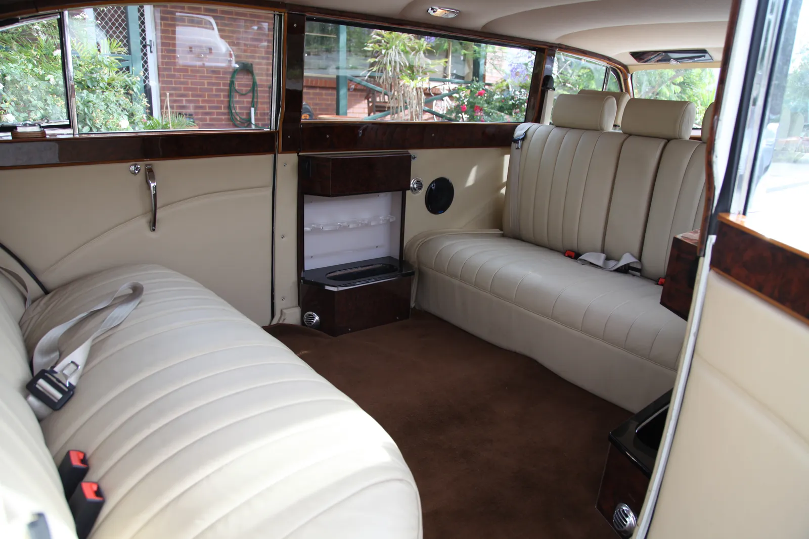 Interior of a luxury vehicle with two cream-colored leather bench seats facing each other, wood trim details, large windows, and a small built-in open bar area with glass holders and a compartment.