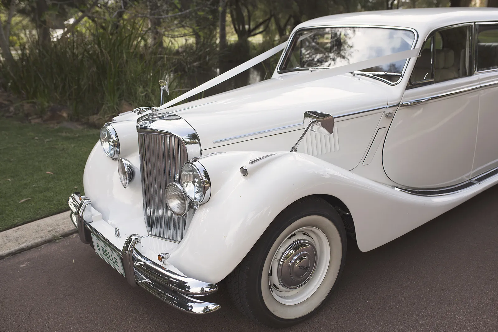 Front side view of a vintage white stretch limousine with chrome details, including a large grille, dual headlights, and a wide front bumper, parked on a paved area beside grass and trees.
