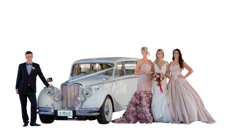 Three women wearing elegant evening gowns and a man in a tuxedo standing next to a classic silver vintage car with the license plate reading '6 BELLE'.