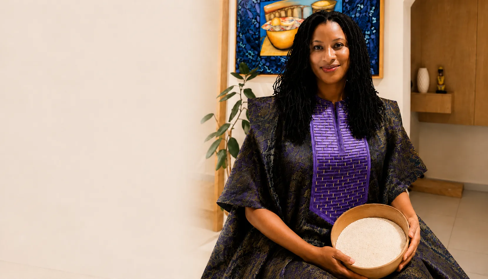 Smiling woman in patterned purple dress holding a wooden bowl filled with white grains.