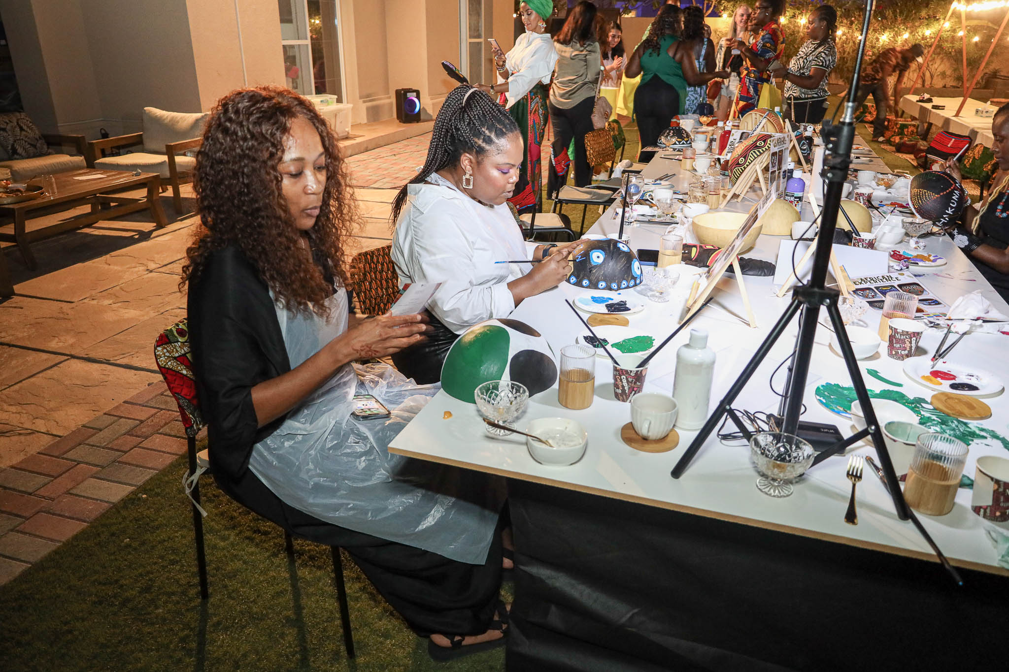 People at a long table painting African-style designs on helmets during an outdoor evening event.