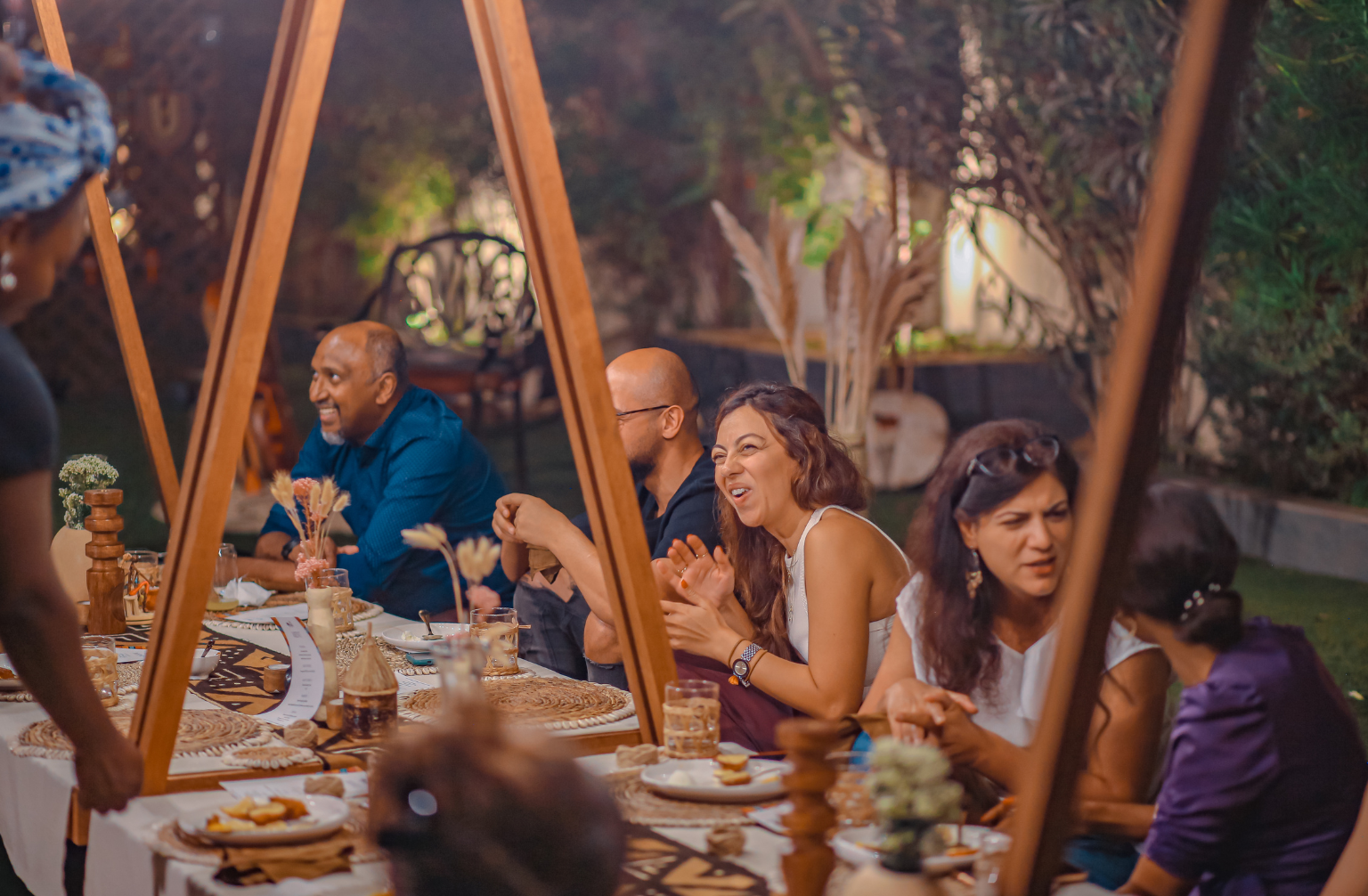 Diverse group dining outdoors at a long table decorated with plants and string lights in a garden.