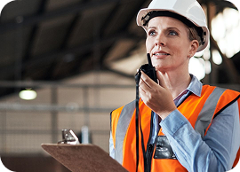 Female construction worker in hard hat and orange vest using walkie-talkie while holding a clipboard.