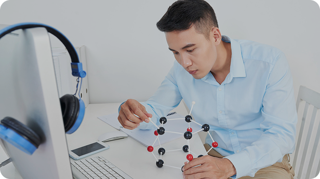 Man in blue shirt studying a molecular model at a desk with computer and phone nearby.