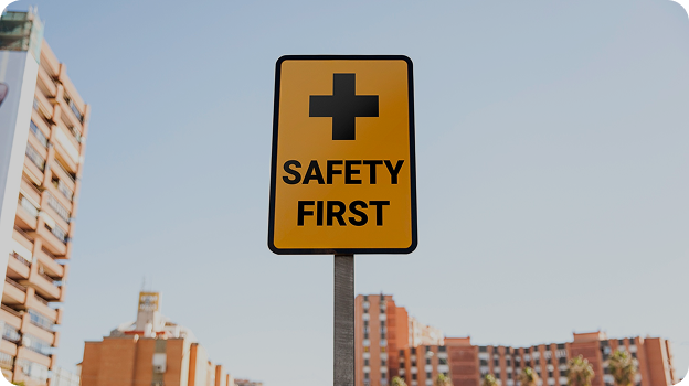 Yellow safety sign with a black cross and text 'SAFETY FIRST' against a cityscape background.