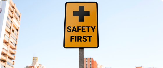 Yellow safety sign with a black cross and text 'SAFETY FIRST' outdoors near buildings under clear sky.