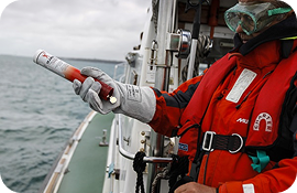 Person in red life jacket and gloves using handheld flare on a boat at sea.