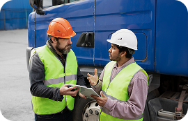 Two construction workers in hard hats and vests discuss work near a blue truck, one holding a tablet.