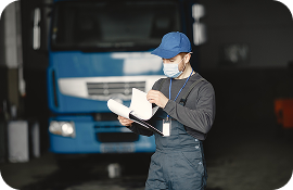 Mechanic in a face mask inspecting paperwork in front of a large blue truck in a garage.
