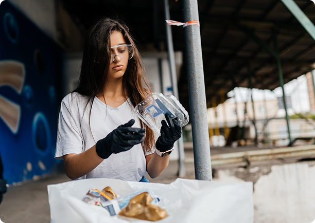 Woman wearing gloves and safety glasses sorting recyclable plastic near a dumpster outdoors.
