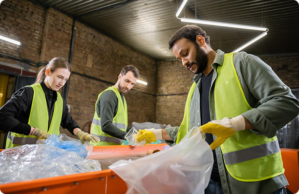 Three workers in safety vests sorting recyclable materials on an orange conveyor belt indoors.