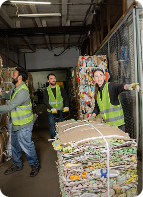 Three workers in safety vests handling large compressed bundles of recyclable materials in a warehouse.