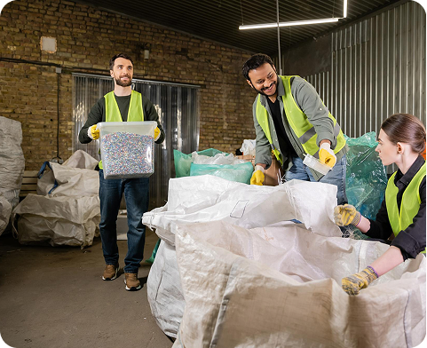 Three workers in safety vests sorting plastic pellets and scraps in large white bags inside a warehouse.