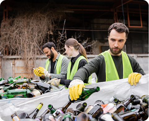 Three workers wearing yellow safety vests and gloves sorting glass bottles for recycling outdoors.