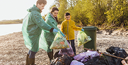 Three people wearing gloves and jackets cleaning up trash near a lake with bags of collected waste