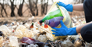 Gloved hands holding a bag, sorting recyclable bottles amid mixed trash outdoors.