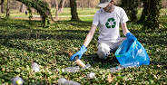 Person wearing mask collects trash outdoors into a blue bag with recycling symbol on shirt.
