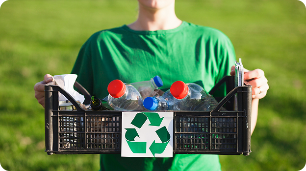 Person in green shirt holding a recycling bin filled with plastic bottles and trash outdoors.
