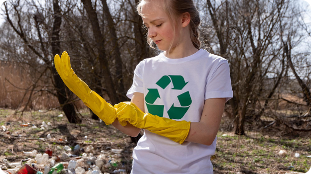 Young girl wearing a white recycling shirt putting on yellow gloves near a littered forest area.