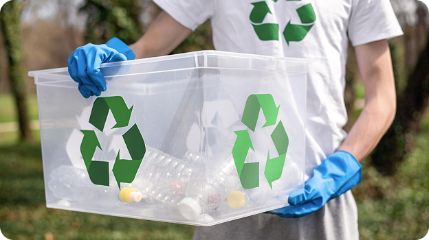 Person wearing blue gloves holding a clear recycling bin with plastic bottles outdoors.