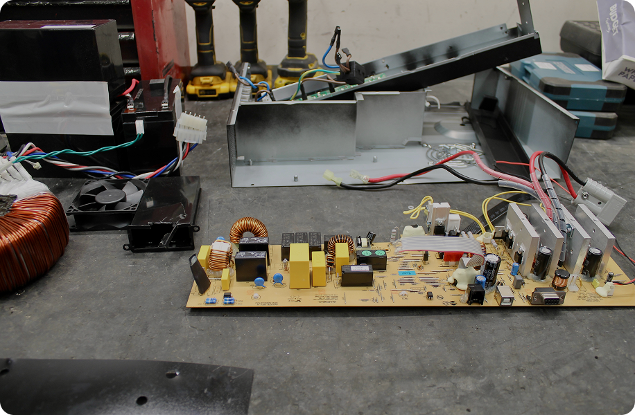 Disassembled electronic circuit board with coils, capacitors, and wires on a workbench with tools in background.
