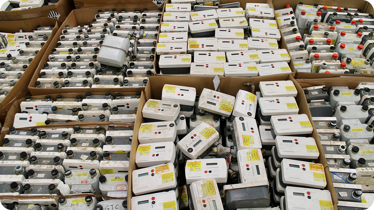 Multiple stacks of utility meters packed tightly in large cardboard boxes on a warehouse floor.