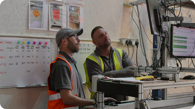 Two men in safety vests looking at a computer screen in a workshop with a whiteboard behind them.