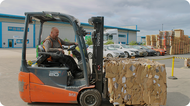 Man operating an orange Toyota forklift moving a large stack of flattened cardboard boxes outside a warehouse.