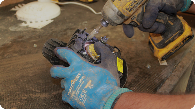 Hands wearing gloves using a power drill to repair a car headlight on a wooden surface.