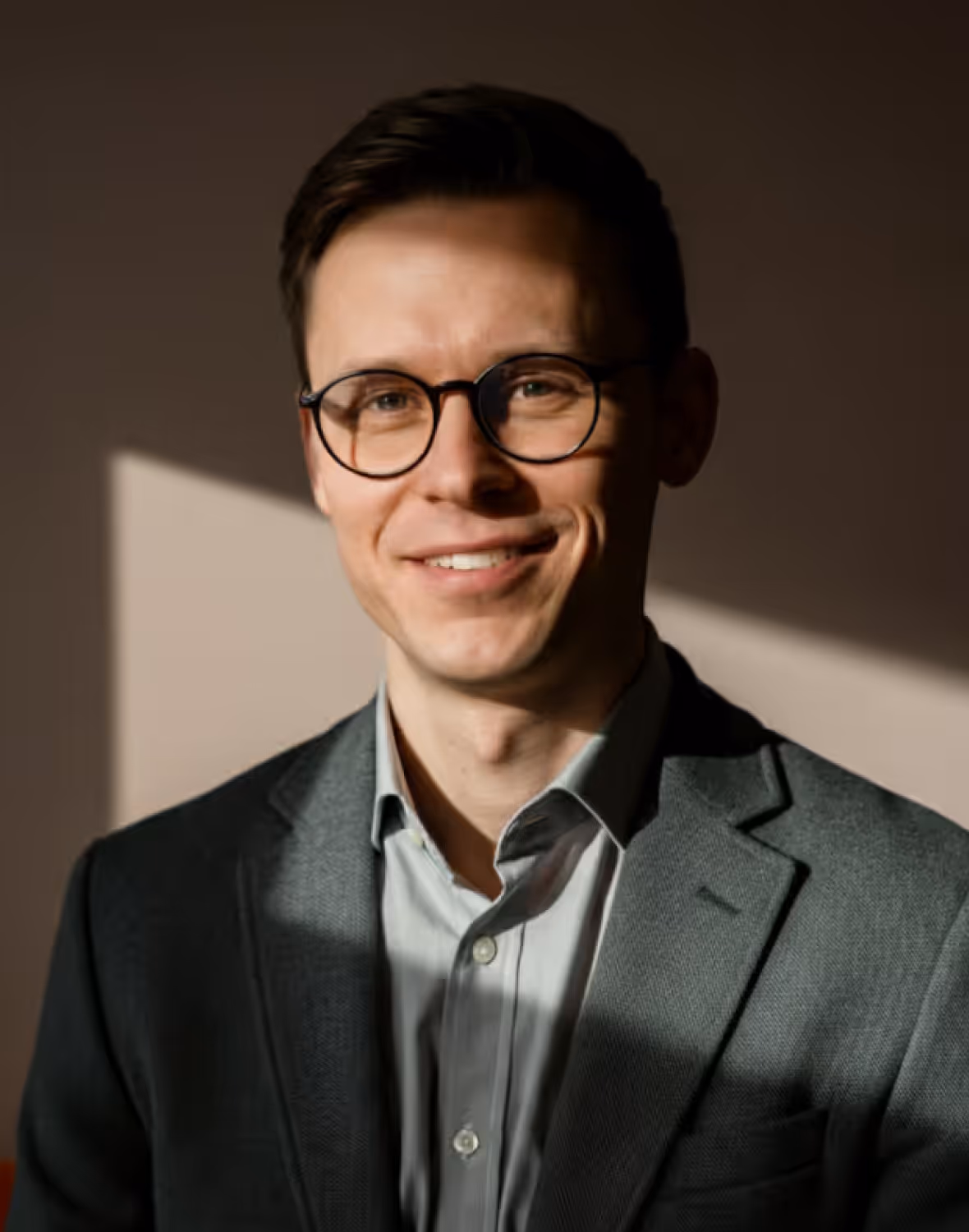 Smiling man with glasses wearing a gray shirt and dark blazer against a shadowed background.