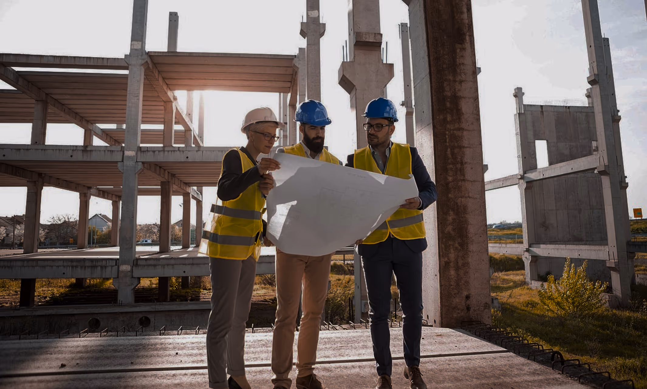 Three construction workers in hard hats and yellow vests reviewing blueprints on a building site.