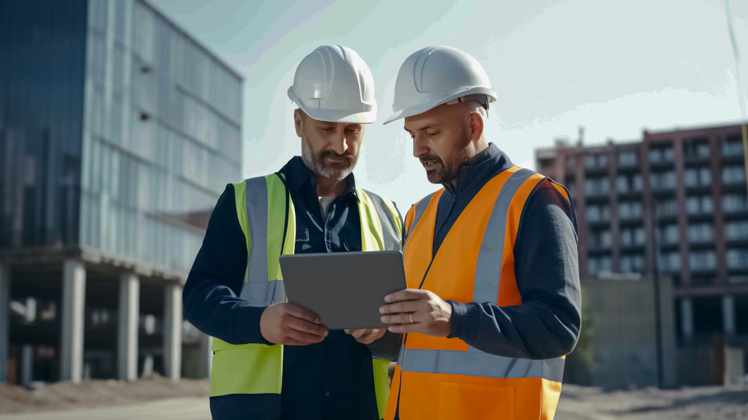 Two construction workers in safety vests and helmets looking at a tablet at a building site.