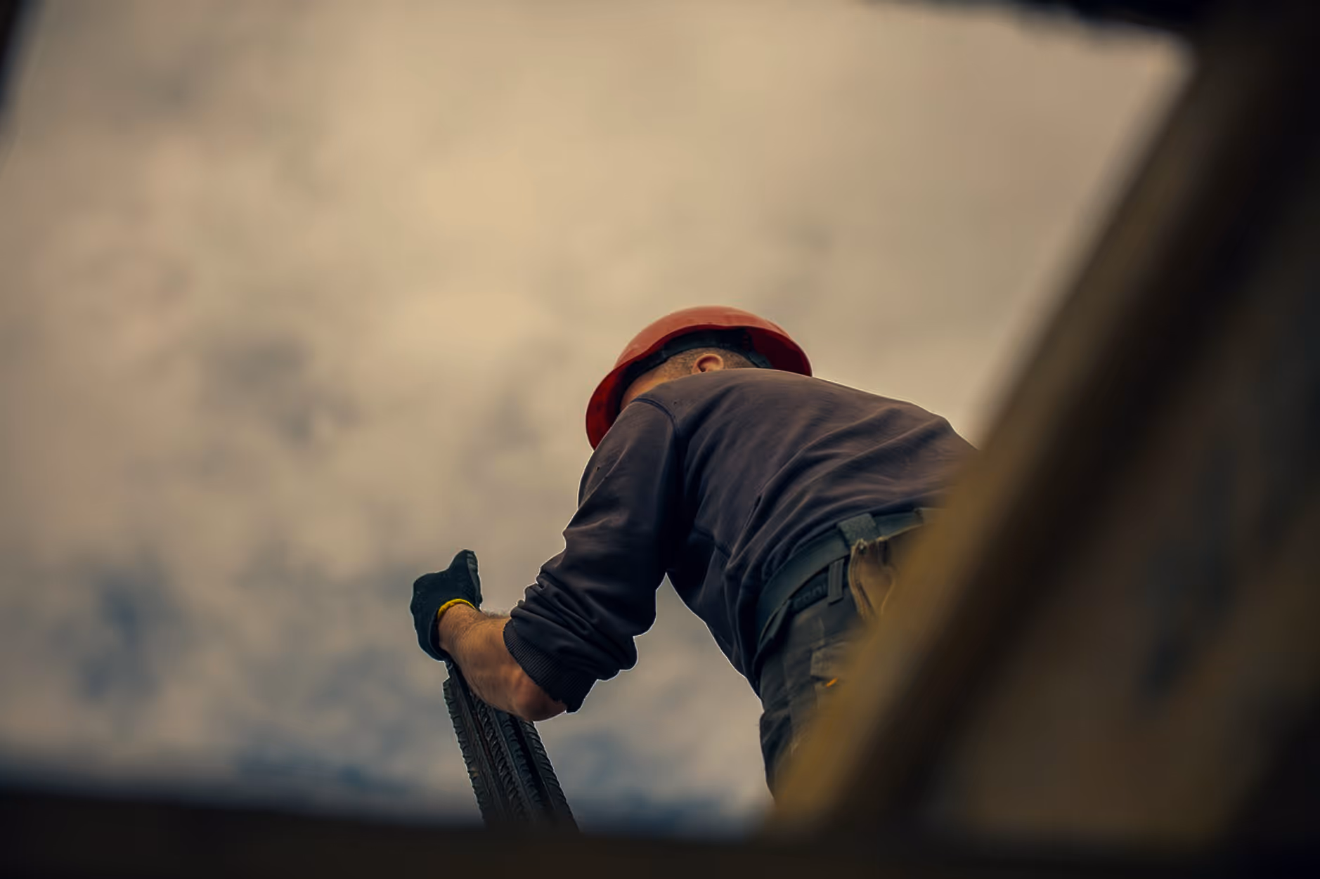 Worker wearing a red hard hat and gloves, holding a tire, viewed from below with sky background.